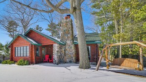 A log cabin with a stone chimney, a swing set, and a red chair outside. - Waterfront Cottage: Family Getaway at Lake Huron! (East Tawas)