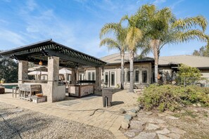 A covered outdoor area with a grill, seating, and a stone pathway leading to a house with a pool.