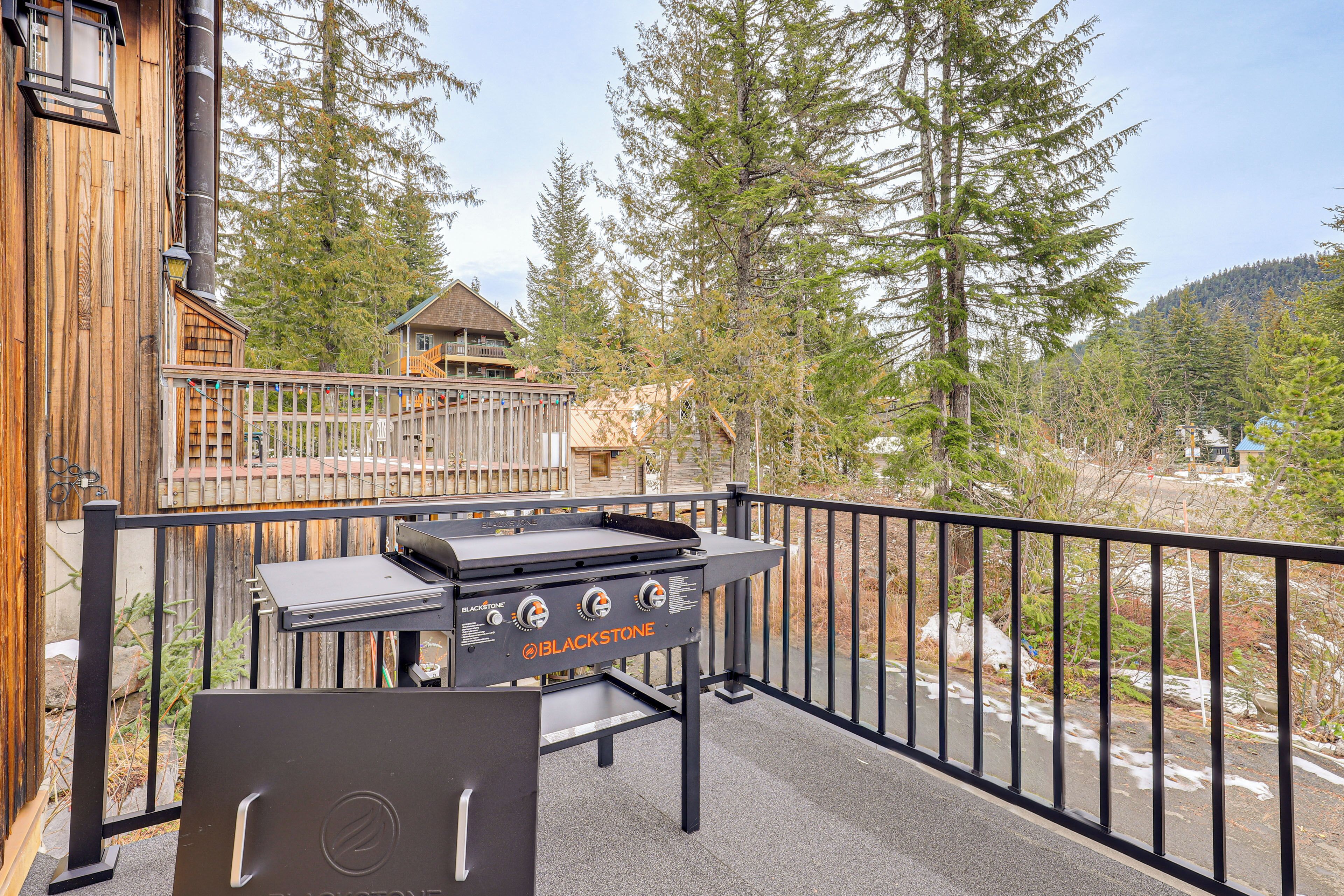 A Blackstone grill on a balcony with a view of a wooden building and surrounding trees.