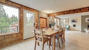 A spacious dining area with a large wooden table and chairs, a stone accent wall, and large windows offering a view of the outdoors. - Après & Adventure: Mt Hood Retreat in Gov Camp! (Government Camp)