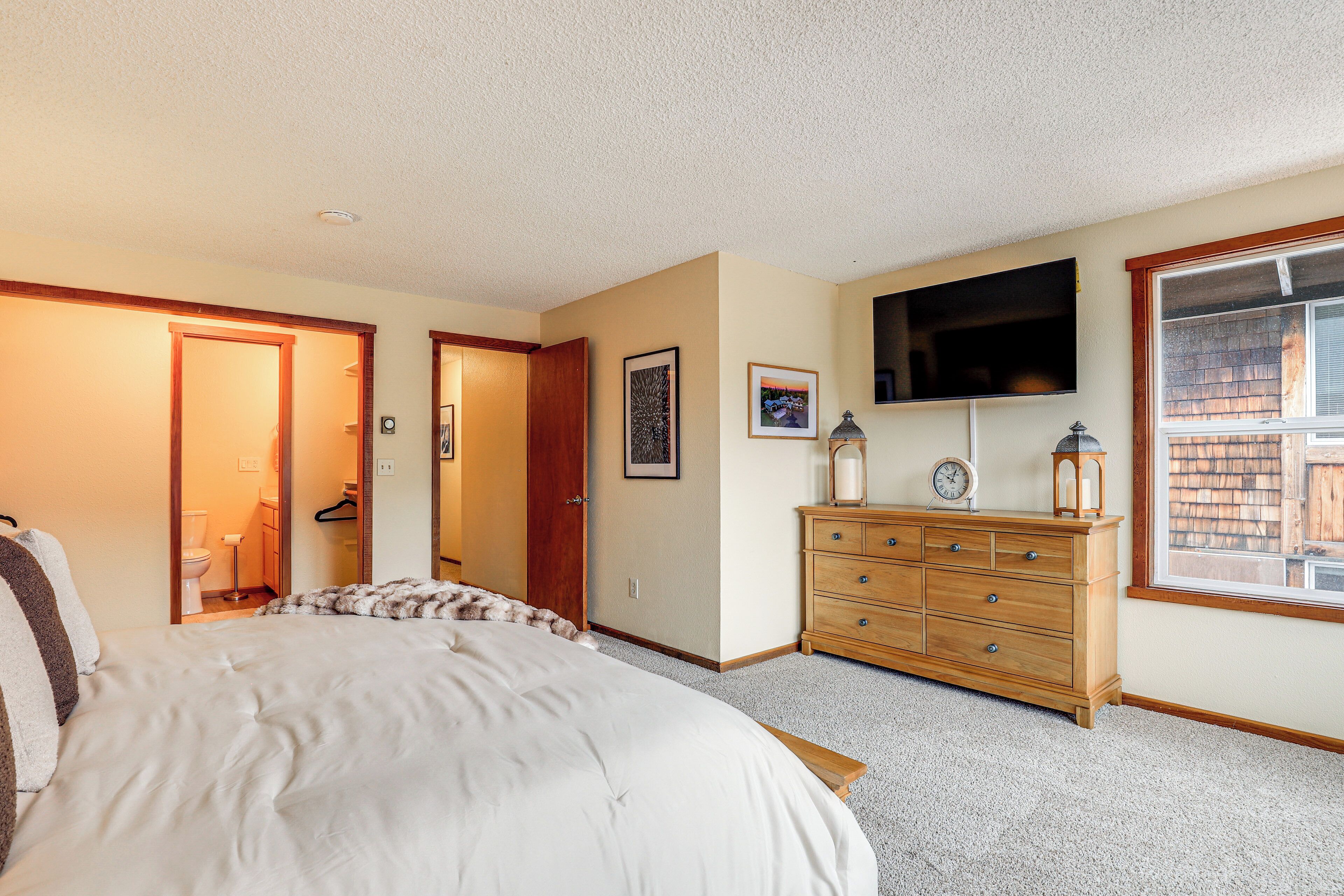 A bedroom with a large bed, a wooden dresser, a flat-screen TV, and a window with a view of a wooden structure outside.