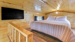 Un dormitorio de cabaña de madera con una cama, un televisor montado en la pared y una ventana con cortinas.