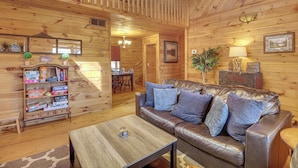 A cozy cabin interior with a leather sofa, wooden walls, a dining area, and a bookshelf. - Hot Tub + Mtn Views: Peaceful Blue Ridge Retreat! (Blue Ridge)