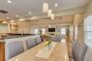 A spacious kitchen with a central island, a dining area with a marble table, and a living area with a TV and artwork. - Saltwater Pool! Epic Galveston Bay Group Getaway! (Dickinson)