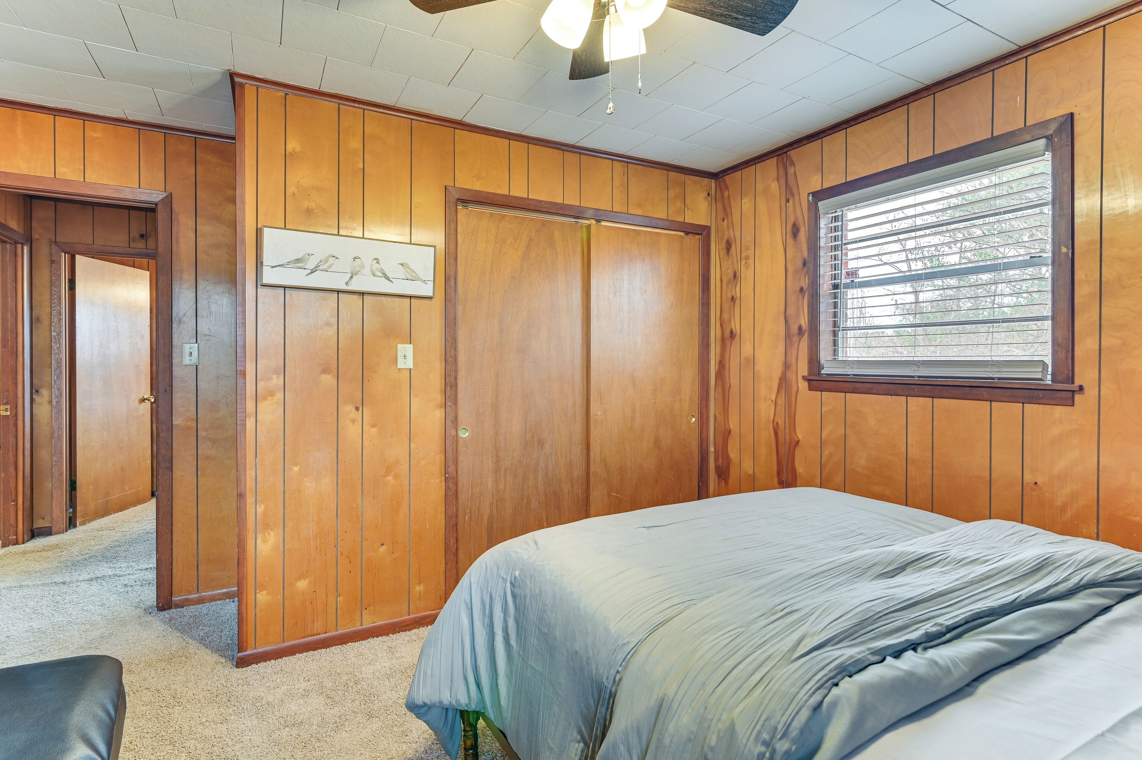 A bedroom with wooden paneling, a bed with a blue cover, a ceiling fan, and a window with blinds.