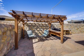 Une pergola en bois, avec deux chaises et une table placée en dessous, installée sur une terrasse en pierre.