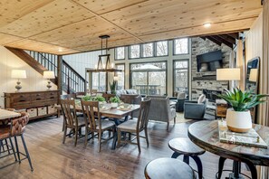 A spacious dining area with a wooden table and chairs, a stone fireplace, and a large window overlooking a deck.