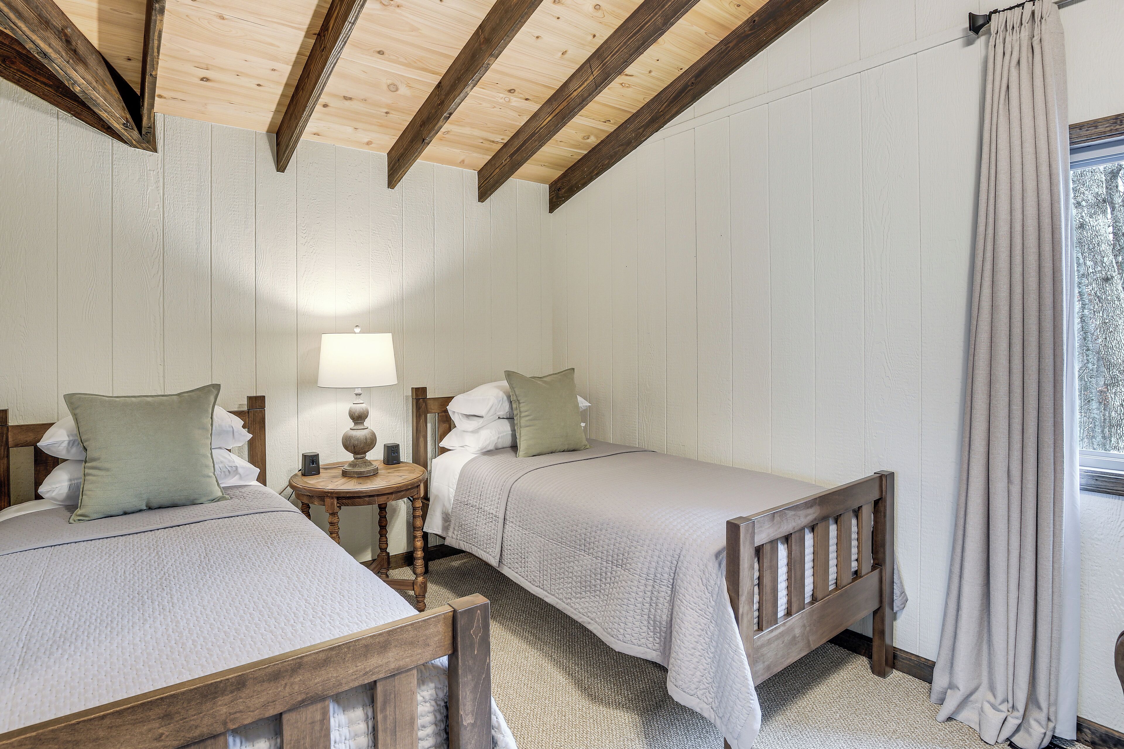 Una habitación con dos camas, cabeceros de madera, una mesita redonda con una lámpara y una ventana con cortinas.