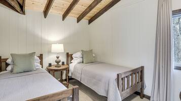 Una habitación con dos camas, cabeceros de madera, una mesita redonda con una lámpara y una ventana con cortinas.