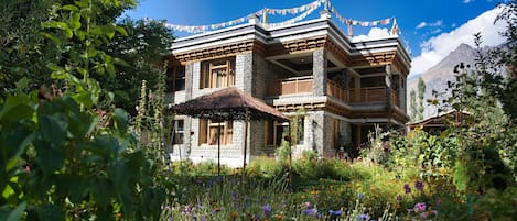 Un edificio tradicional de varios pisos con una estructura de techo de paja al frente, rodeado de un jardín vibrante con diversas flores silvestres.