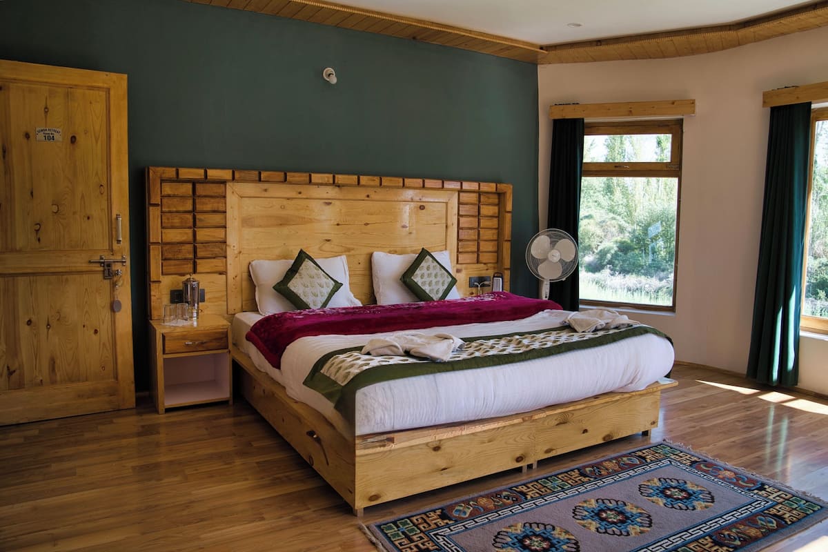 Una cama con estructura de madera, cabecera, mesita de noche, ventilador y vista a la vegetación a través de una ventana.