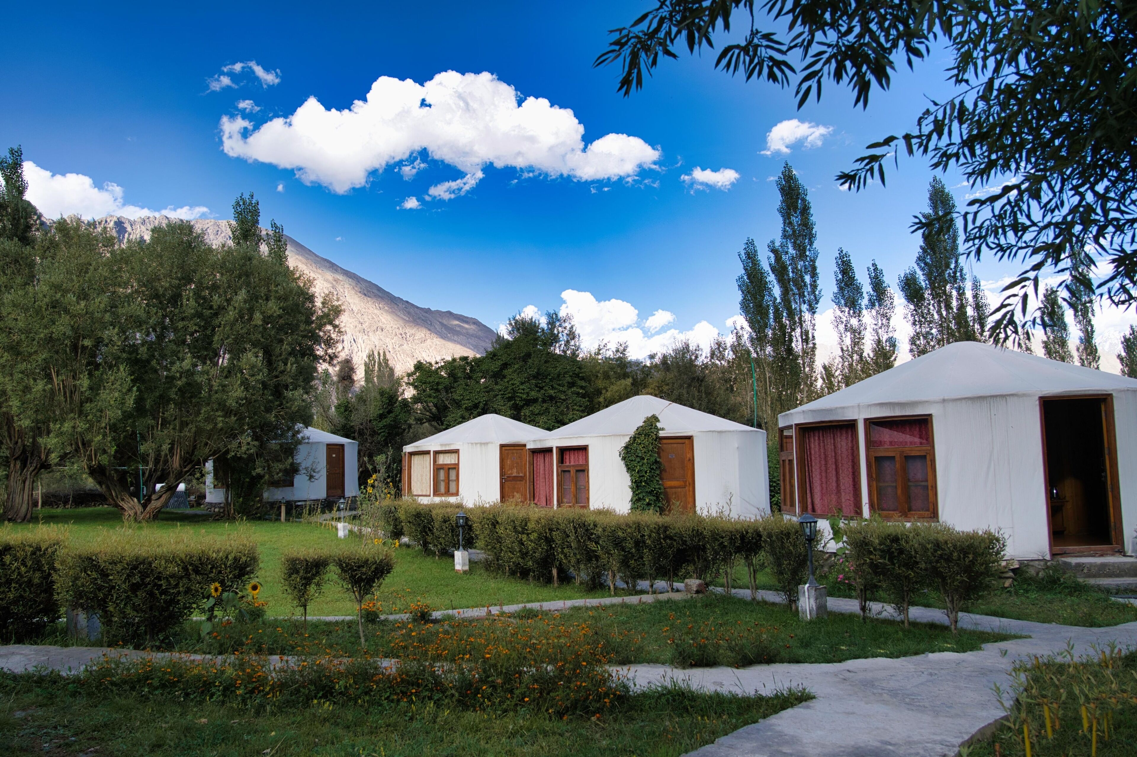A row of white yurts with wooden doors and windows, surrounded by well-maintained hedges and a clear blue sky.