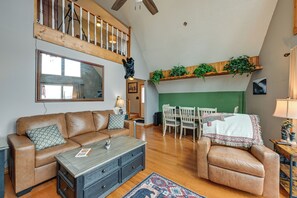 Un salón amplio con techo alto, una escalera de madera, una zona de comedor con mesa y sillas, y una pared verde decorativa.