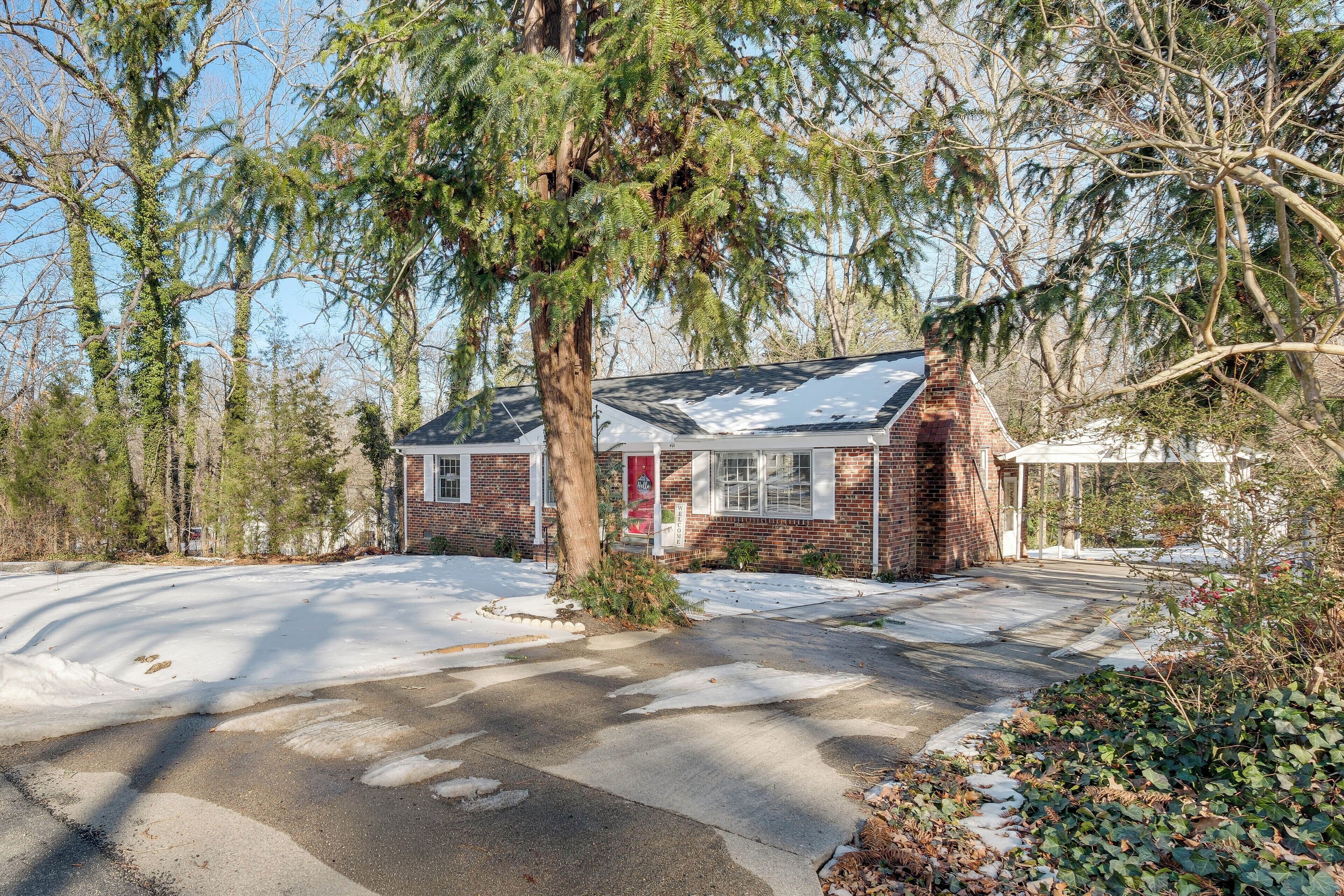 A small brick house with a red door, surrounded by trees and a snow-covered driveway.