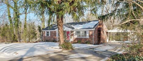 A small brick house with a red door, surrounded by trees and a snow-covered driveway.