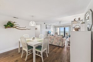 A dining area with a white table and chairs, a sofa, and a ceiling fan. - Castle Reef 524 beautifully furnished, ocean views! (New Smyrna Beach)