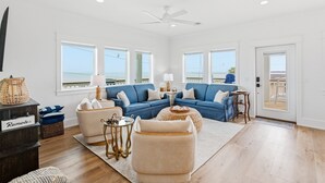 A spacious living room with a large blue sofa, a beige armchair, and a small side table. There are large windows offering a view of the ocean. - Coastal Breeze Cottage near beach water views (Port O'Connor)