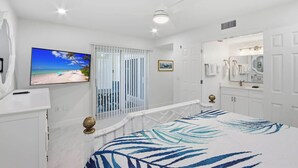 Ein Schlafzimmer mit Bett, Fernseher, einem Balkon mit Blick auf den Strand und einem Badezimmer mit Spiegel und Waschbecken.