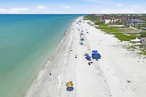 Une vue aérienne d’une plage avec une rangée de parasols, quelques personnes et des bâtiments en arrière-plan.