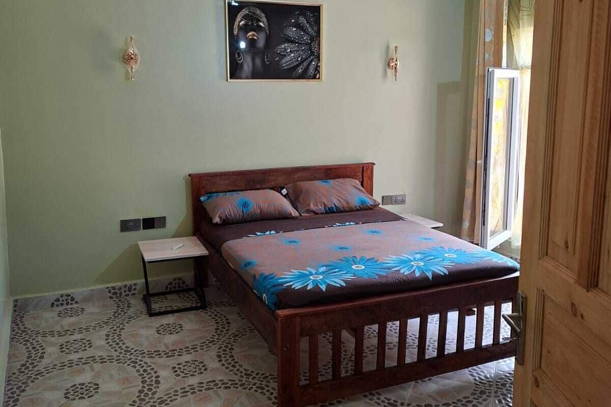 A bedroom with a bed, a nightstand, a ceiling fan, a framed picture on the wall, and patterned floor tiles.