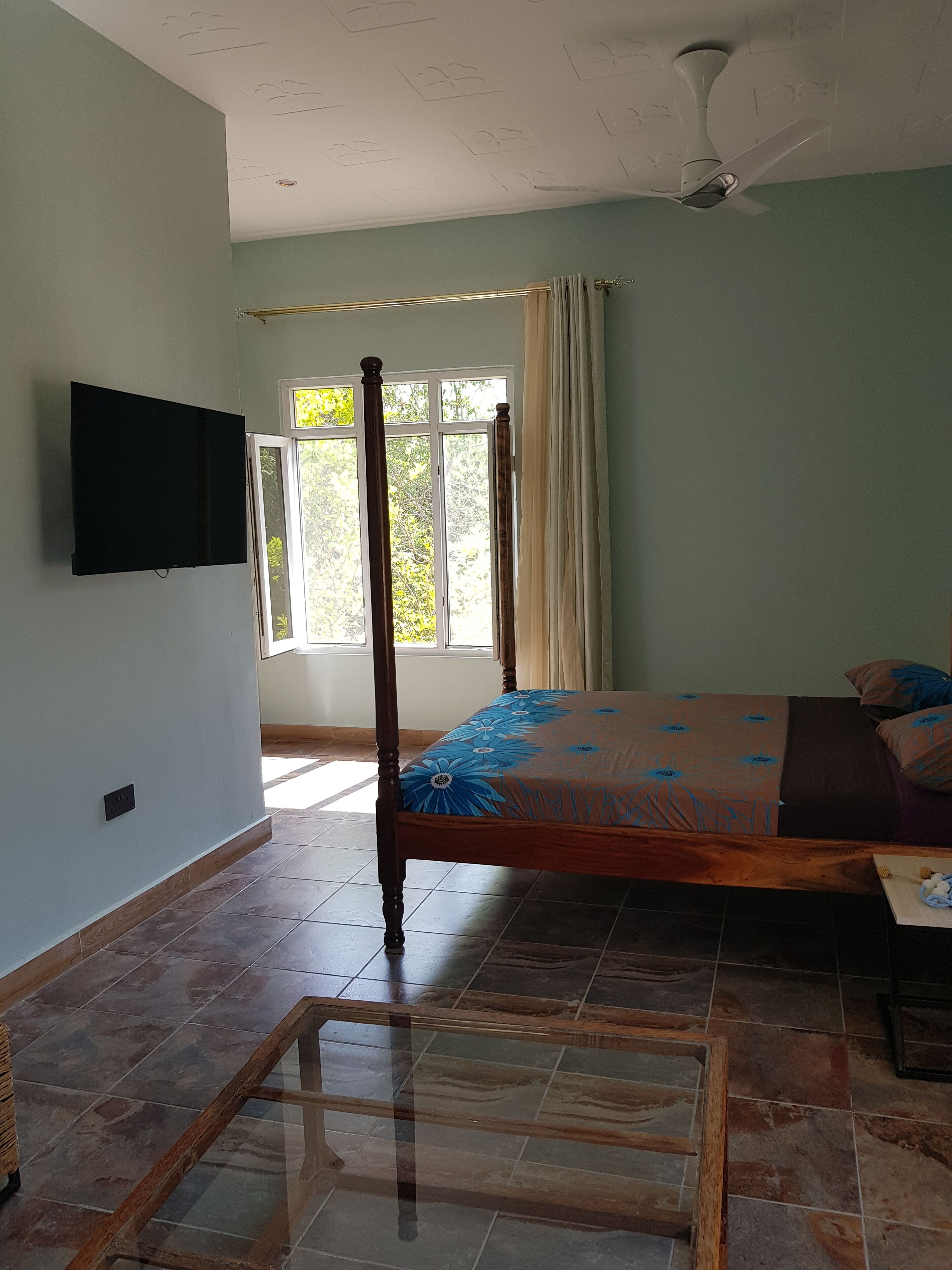 A bedroom with a bed, a ceiling fan, a window with curtains, and a television on the wall.
