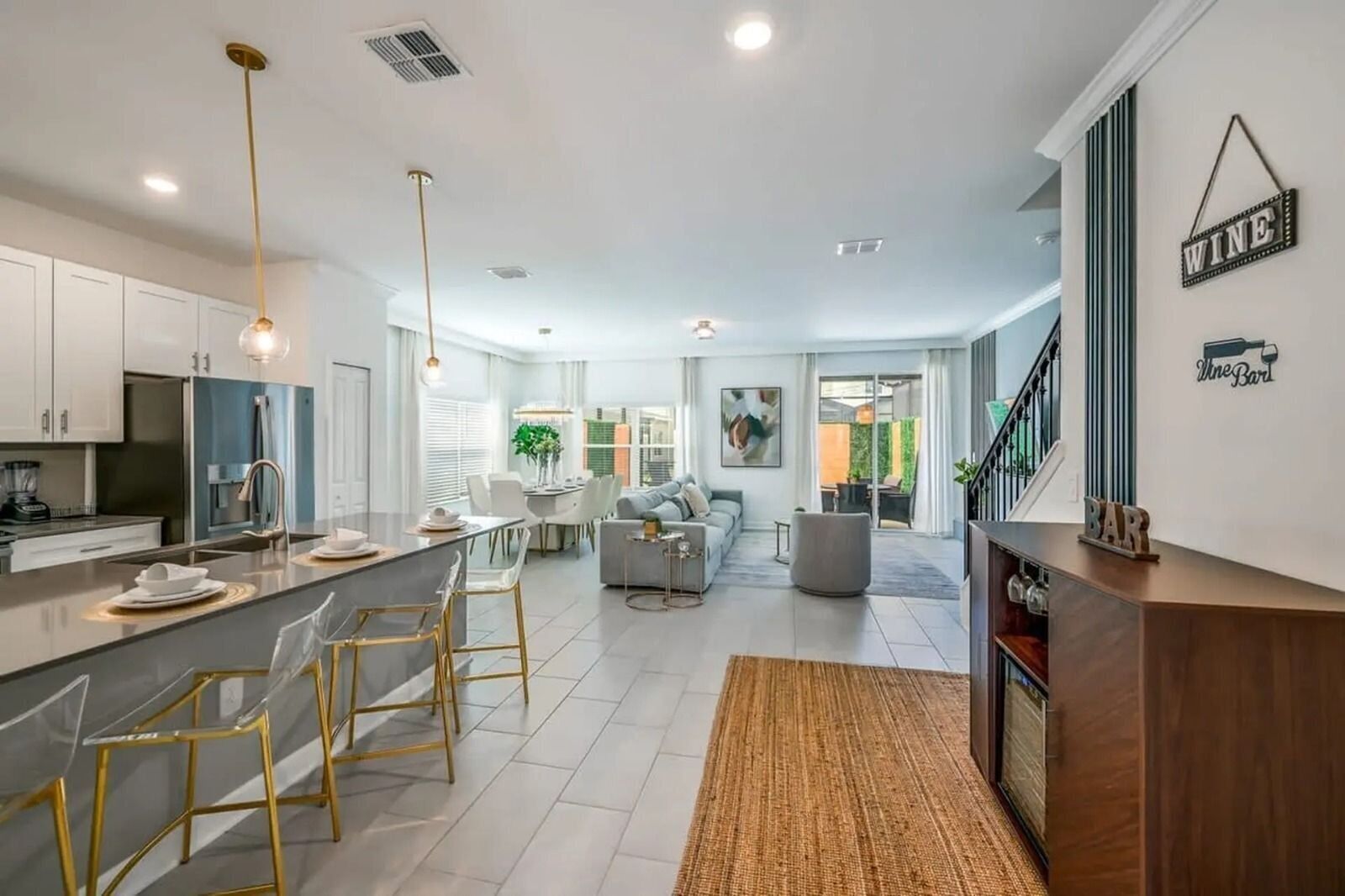 A modern kitchen with a central island, dining area with chairs, a living room with a sofa, and a staircase leading to a bar area.