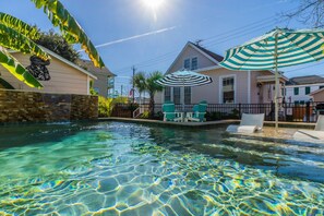 A swimming pool with clear water, surrounded by houses, a stone wall, and a large striped umbrella. - Iggy's Oasis- Relax, Unwind & Recharge (Galveston)