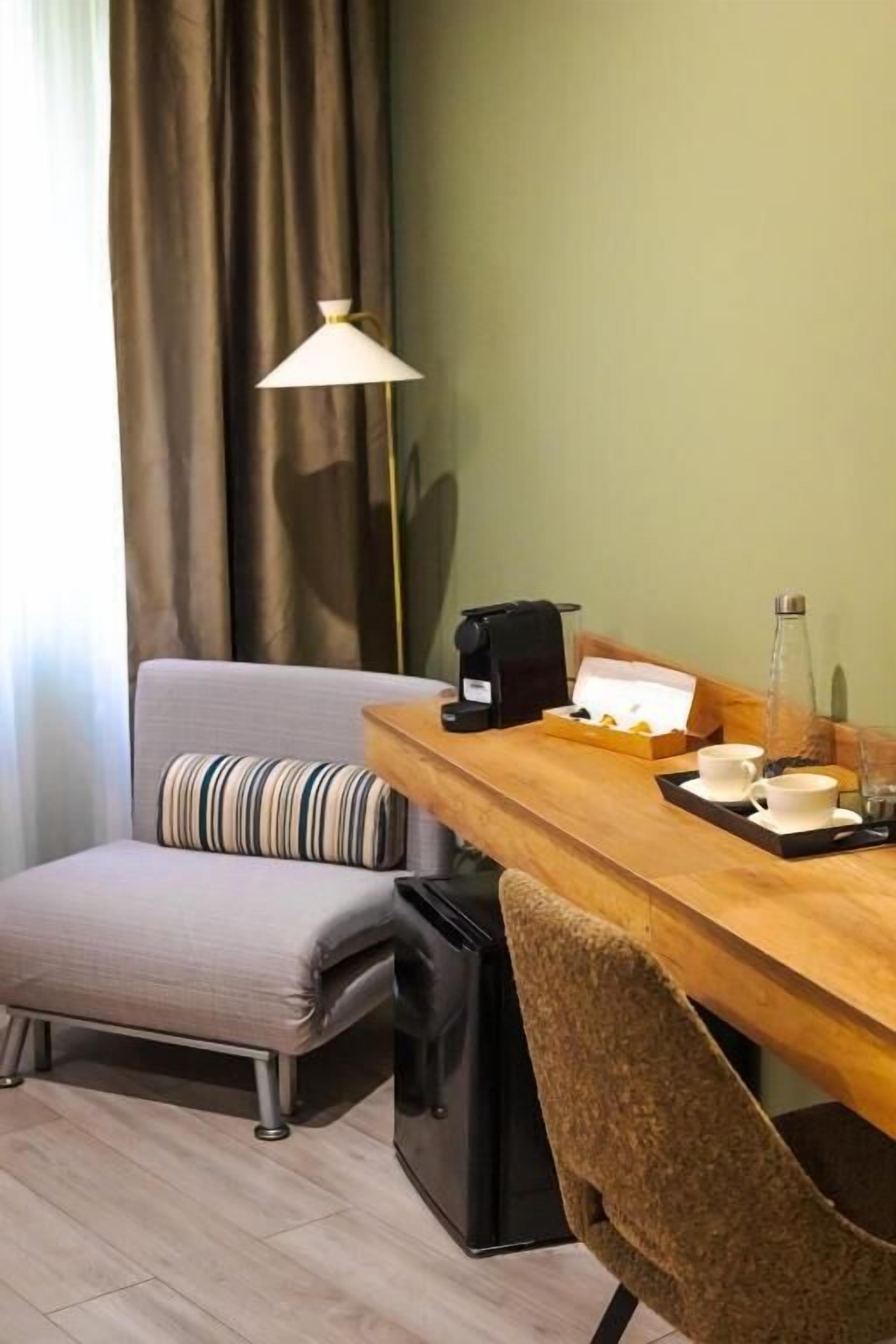 Una habitación pequeña y acogedora con un escritorio de madera, una silla, un sofá, una lámpara y una cafetera.