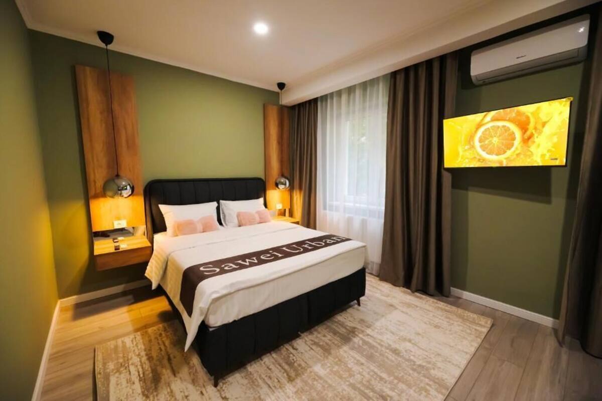 Habitación de hotel con cama, lámparas de noche, televisor y un ventanal con cortinas.