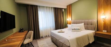 Habitación de hotel con cama, escritorio, silla, televisor y ventana con cortinas.