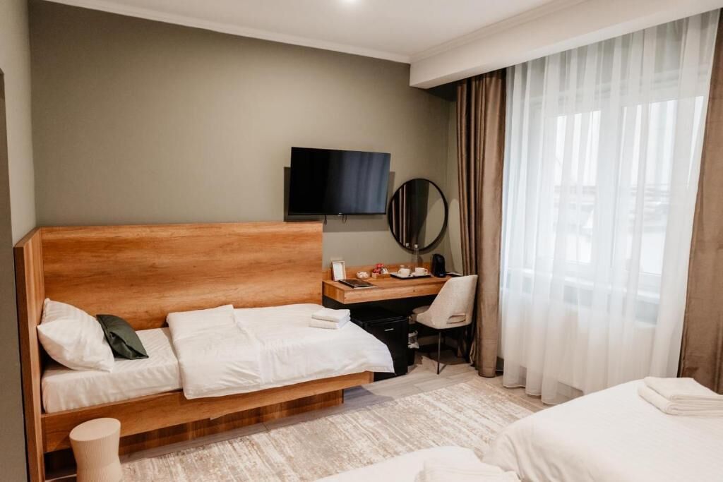A hotel room with a wooden bed, a desk with a chair, a television, and a large window with curtains.