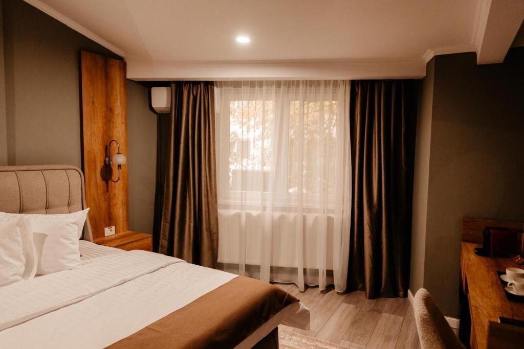 Habitación de hotel con cama, escritorio, silla, ventana con cortinas y lámpara en la pared.