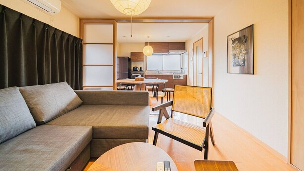 Un salón moderno con un sofá, una mesa de centro de madera y una silla de ratán. Se ve una cocina al fondo con una mesa de comedor y sillas.