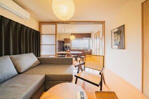 Uma sala de estar moderna com um sofá, uma mesa de centro de madeira e uma poltrona de rattan. Uma cozinha é visível ao fundo, com uma mesa e cadeiras de jantar.