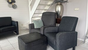 A modern living room with a staircase, a black leather sofa, a grey armchair, and a black ottoman. - For rent Island Happiness! (Oudesluis)