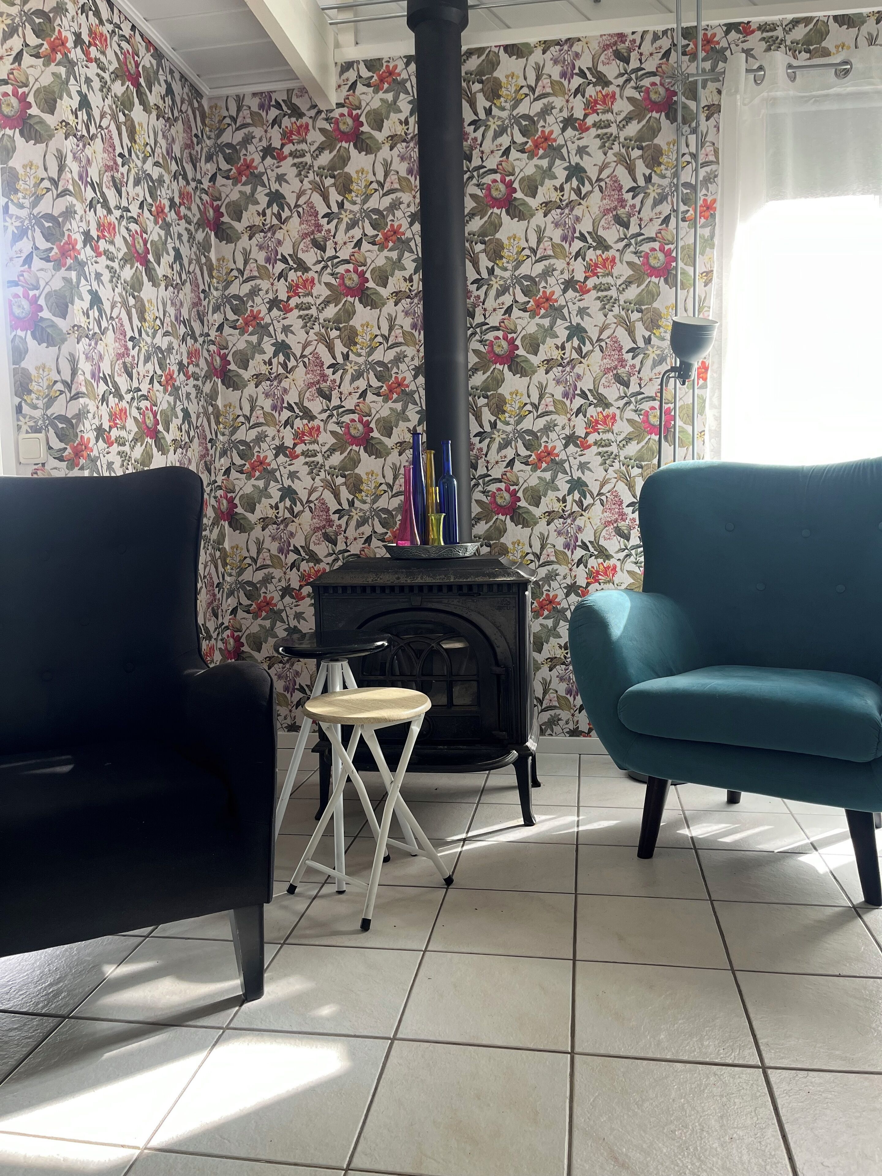 A living room with floral wallpaper, a black fireplace, two chairs, and a small stool.