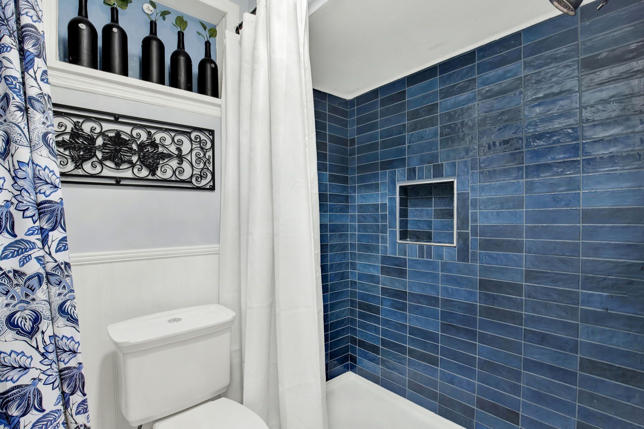 A bathroom with a toilet, a shower area with blue tiles, and a decorative shelf with bottles.