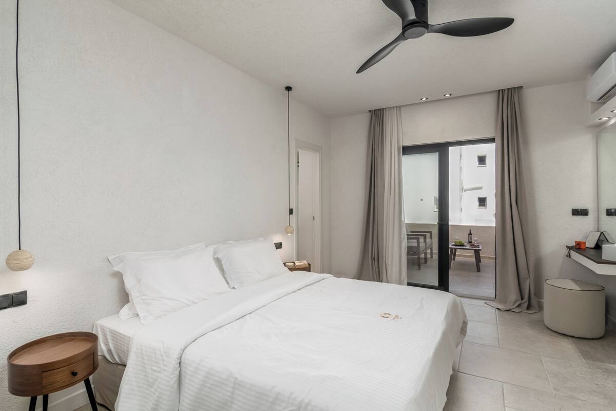 A modern hotel room with a bed, a round wooden nightstand, a ceiling fan, and a view to the outside.