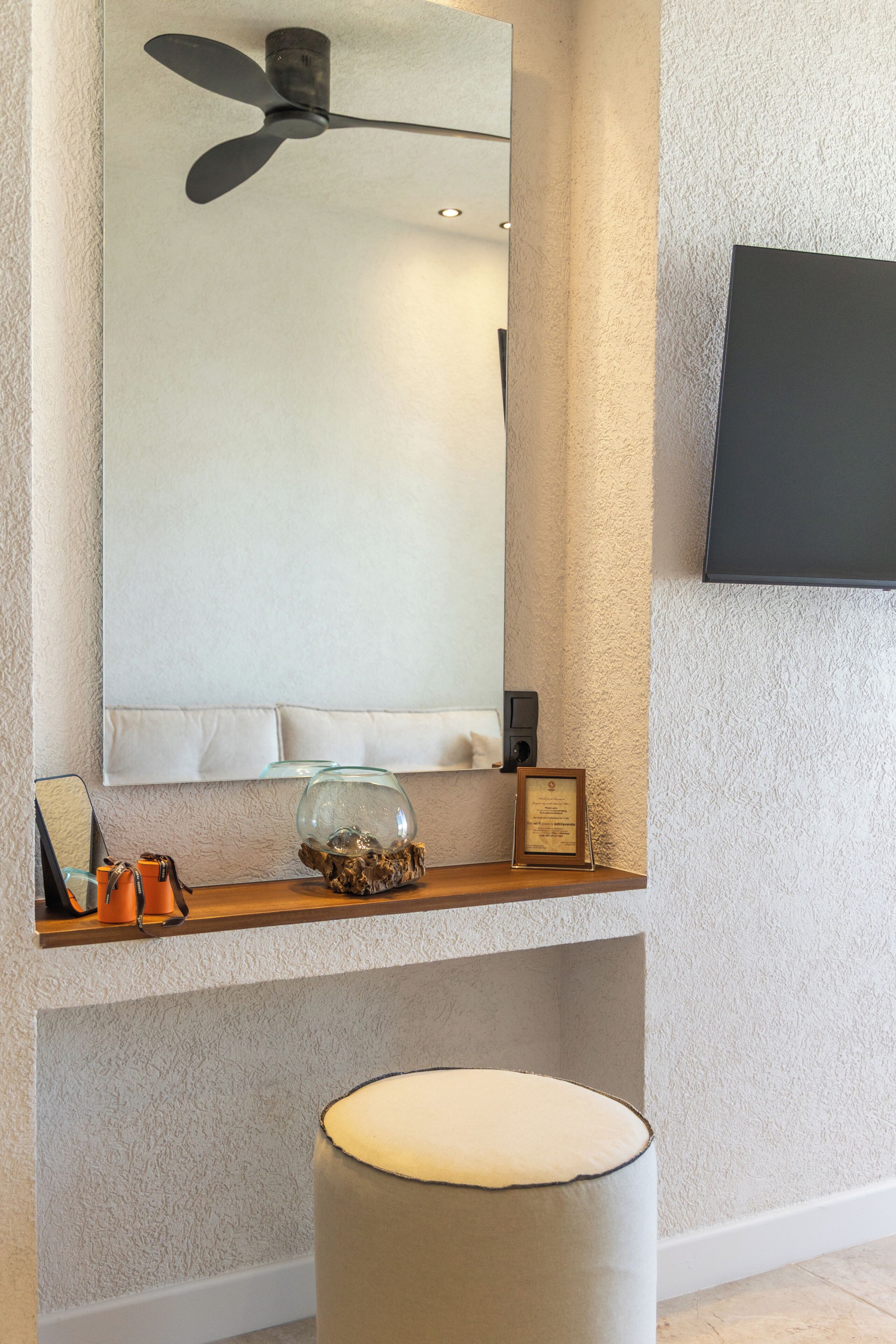 Une pièce dotée d’un ventilateur de plafond, d’un téléviseur à écran plat fixé au mur, d’une étagère décorative avec un saladier en verre et un tableau encadré, ainsi que d’un tabouret rembourré.
