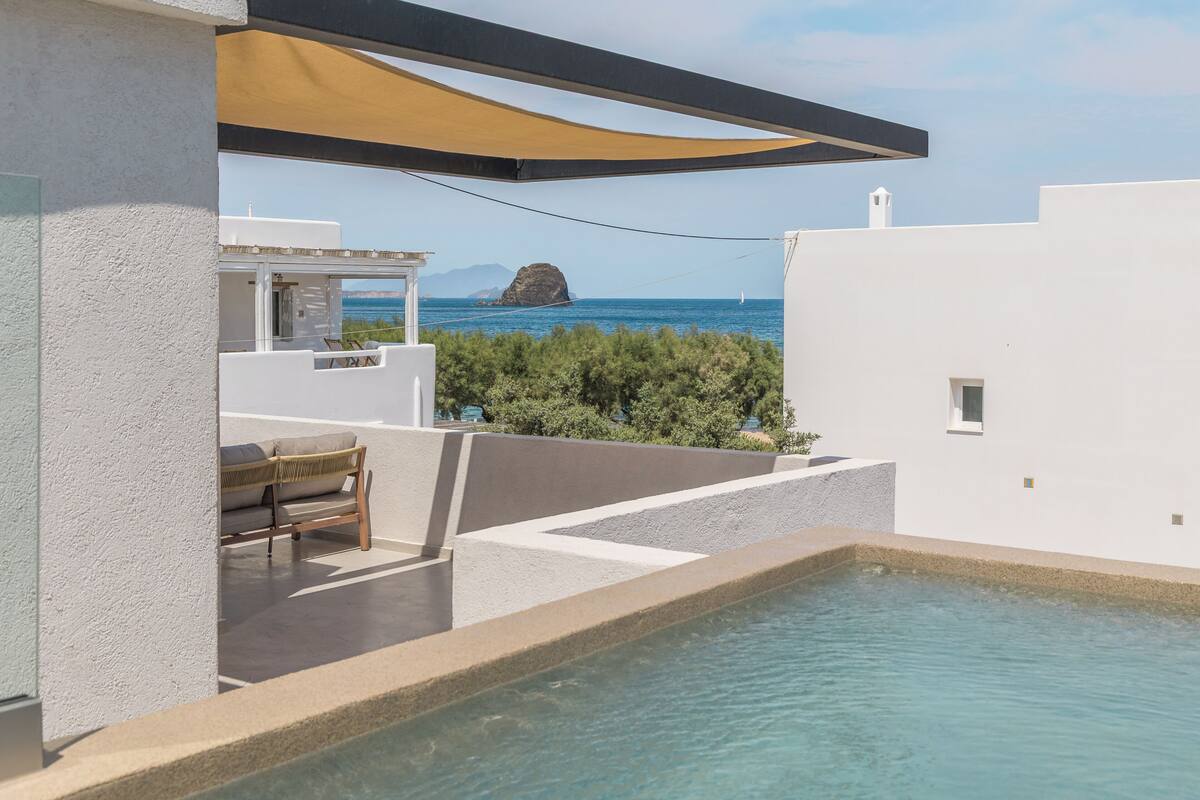 A pool area with a view of the sea, a white building, and a shaded seating area.