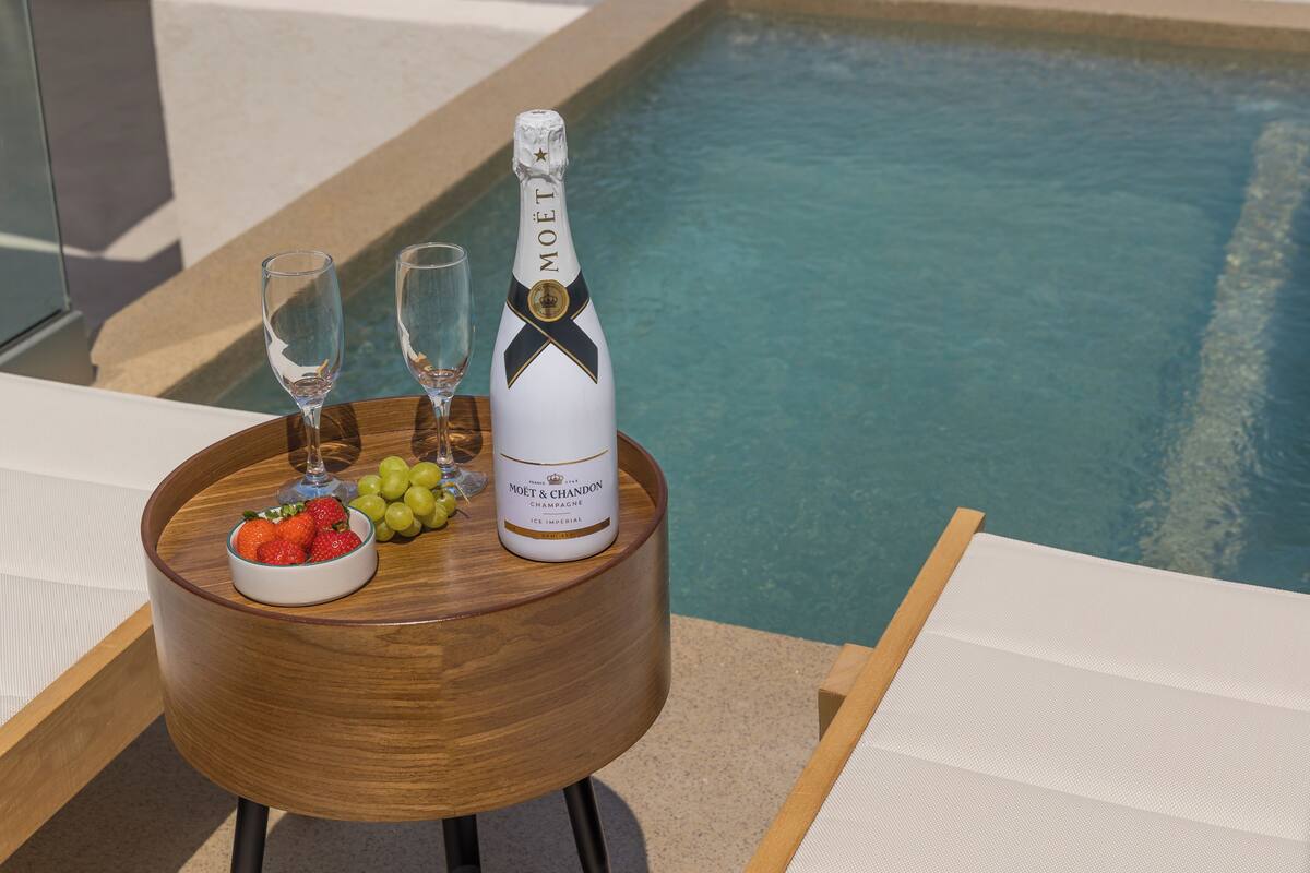 A wooden table with a bottle of Moët & Chandon champagne, two glasses, and a bowl of fruit by a poolside.