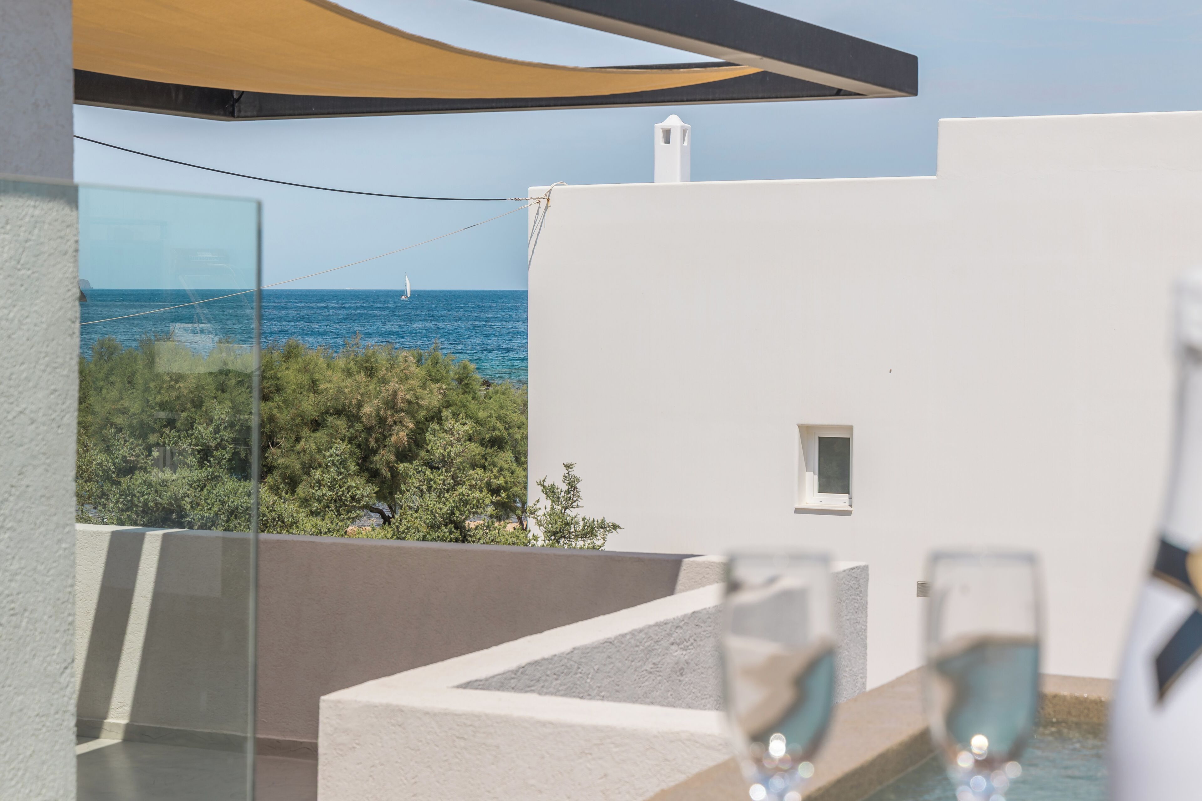 Un balcón con vista al mar, un edificio blanco y una mesa de vidrio con copas de vino.