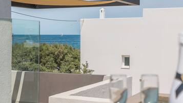 A balcony with a view of the sea, a white building, and a glass table with wine glasses.