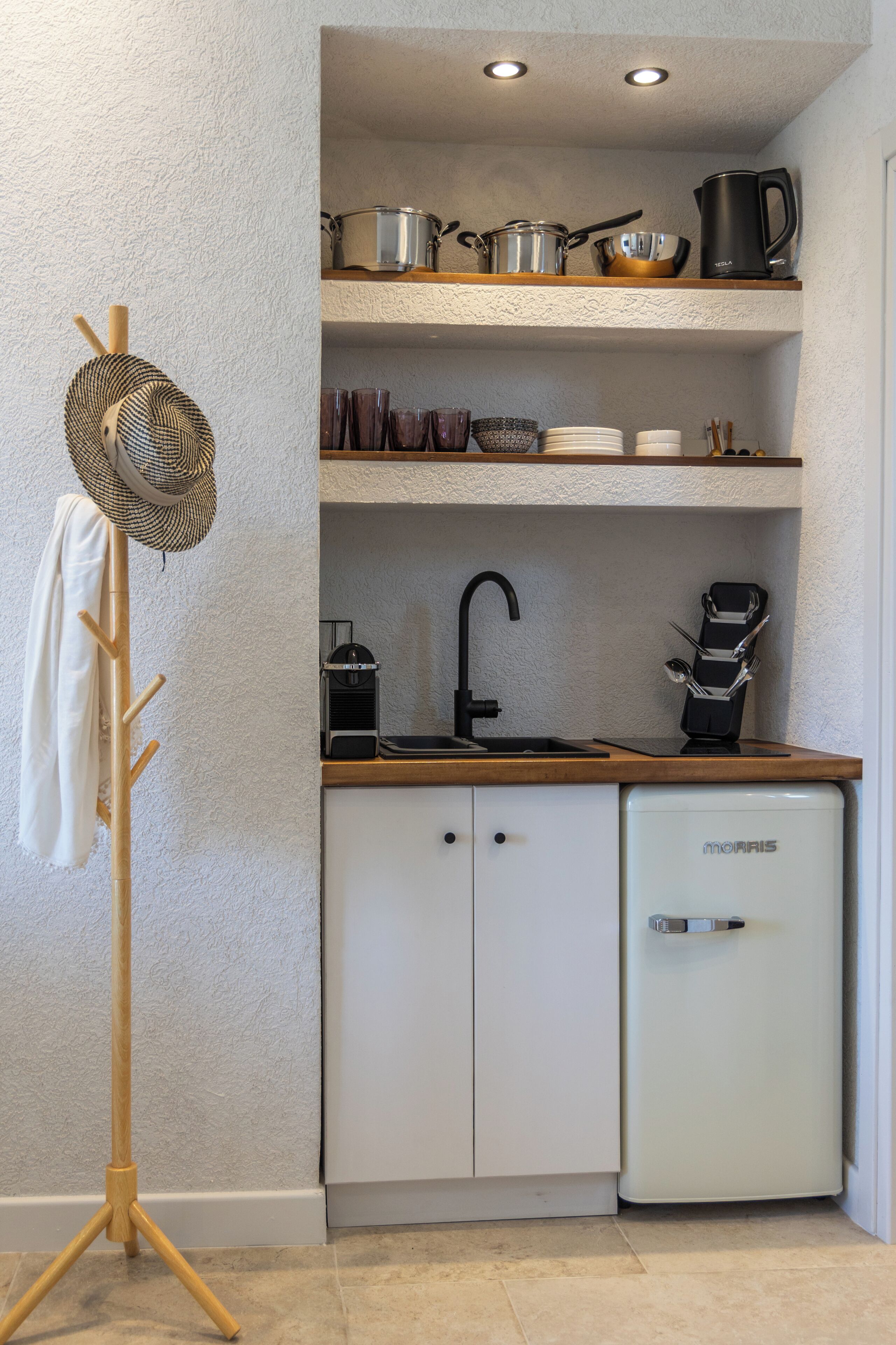 A compact kitchen with white cabinets, a small refrigerator, a coffee maker, and a shelf with pots and a kettle.