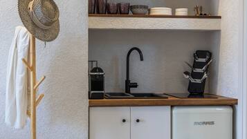 A compact kitchen with white cabinets, a small refrigerator, a coffee maker, and a shelf with pots and a kettle.