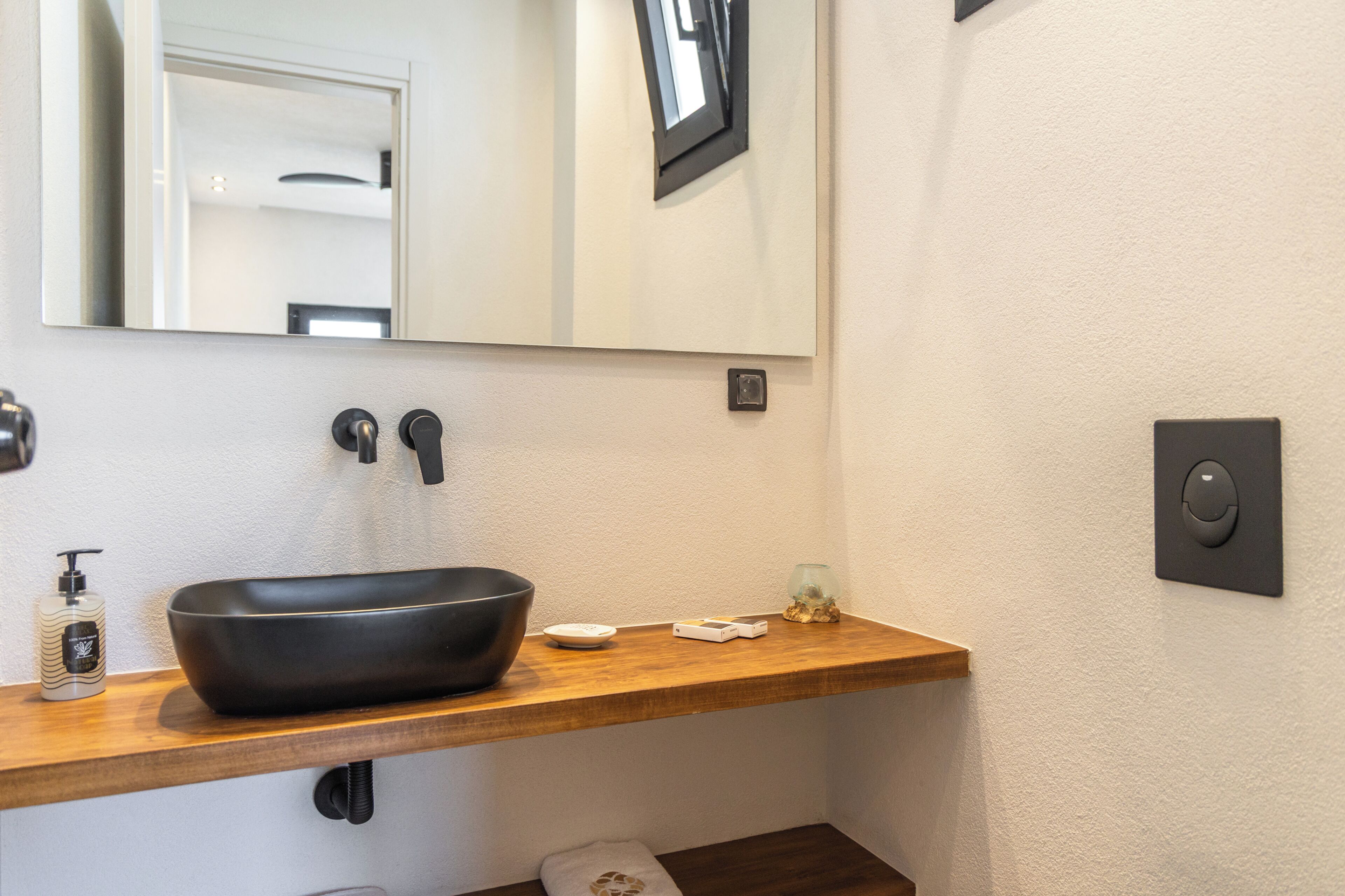 Un baño con un lavabo negro tipo recipiente, un espejo, una toalla, un dispensador de jabón y un interruptor de luz.