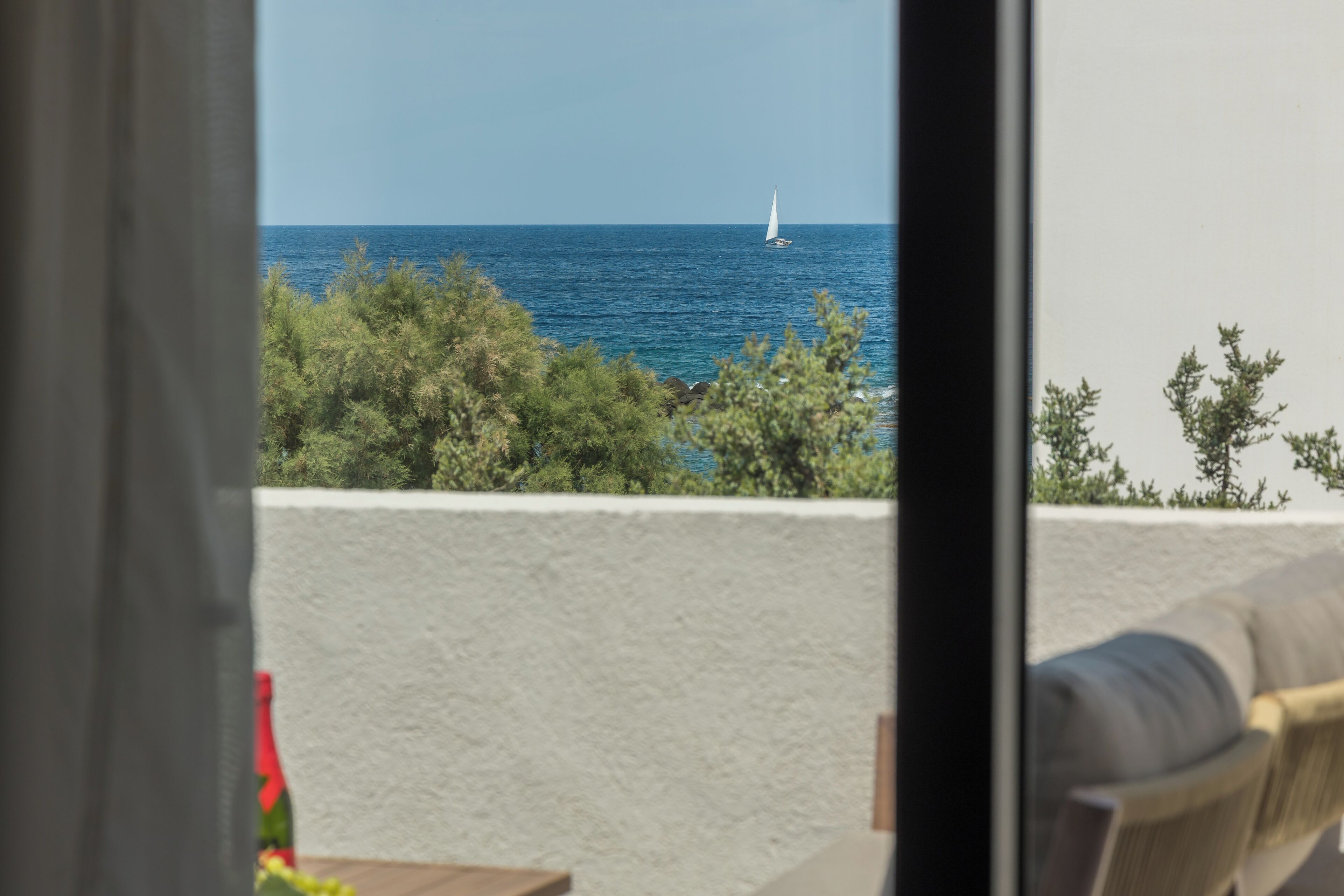 Vista al mar desde un balcón con un velero, vegetación y una pared blanca.