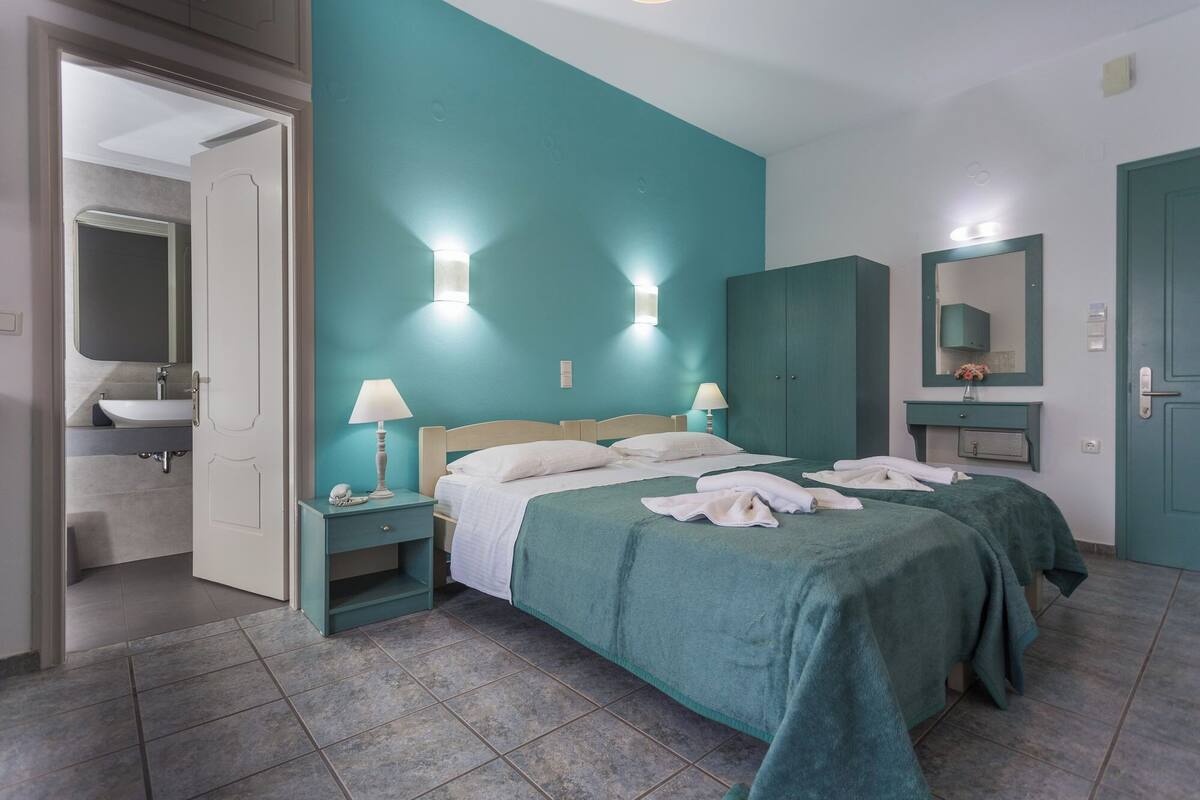 A hotel room with teal walls, a bed with white linens, two bedside tables with lamps, a large mirror, and a bathroom visible through an open door.