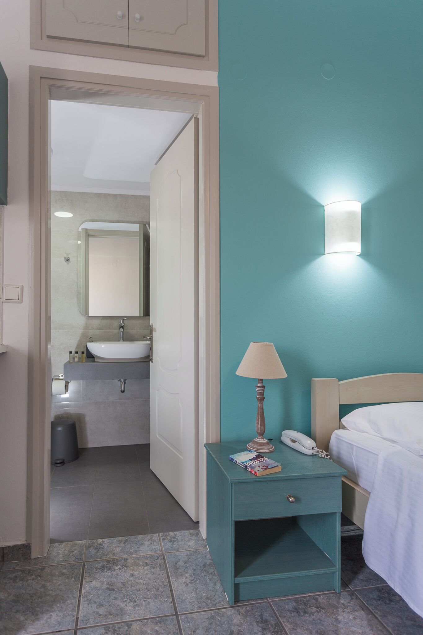 A room with a teal wall, a white bed, a nightstand with a lamp, a phone, and a book. There is a door leading to a bathroom with a sink and mirror.