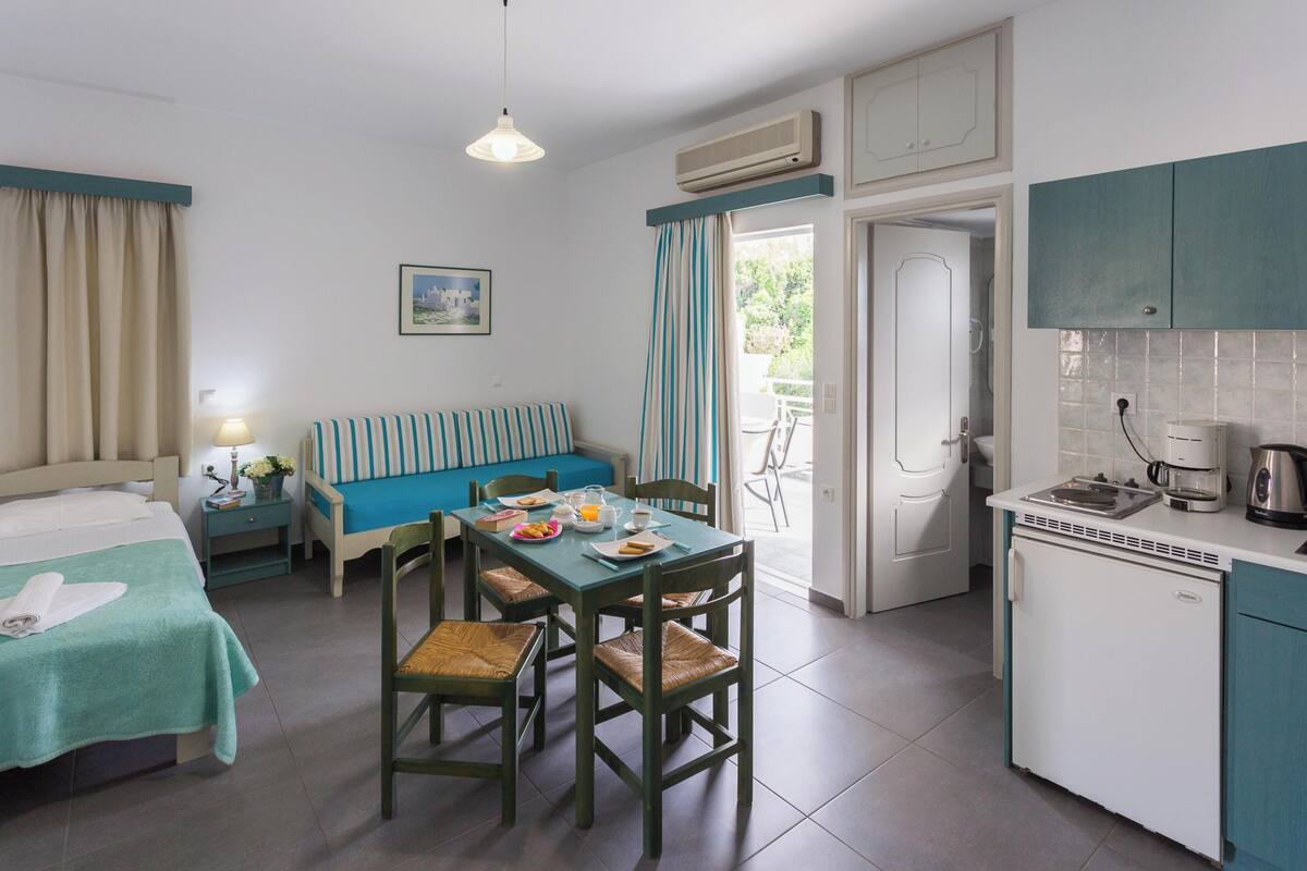 A room with a bed, a dining table with chairs, a kitchenette, and a balcony with curtains.
